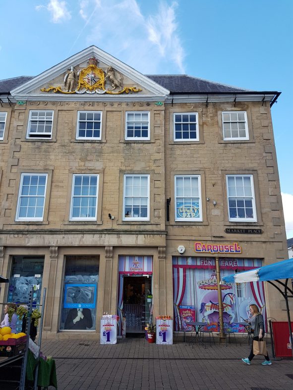 Frontal view of imposing 18th century three storey building in ashlar stone with rows of seven sash windows on the first and second floor. It has a 20th century timber shop front, coursed stone above and a Welsh slate roof with a gilded coat of arms on the pediment.