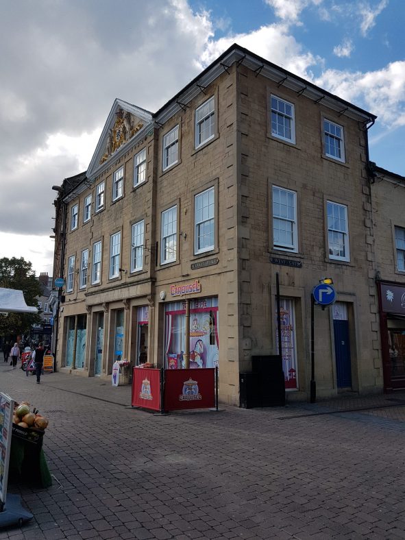 Corner view of an imposing 18th century three storey building in ashlar stone with rows of seven sash windows on the first and second floor. It has a 20th century timber shop front, coursed stone above and a Welsh slate roof with a gilded coat of arms on the pediment.